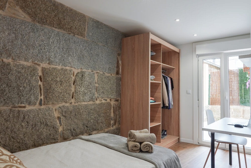 Habitación moderna con pared de piedra, cama doble, armario abierto y escritorio frente a la ventana.